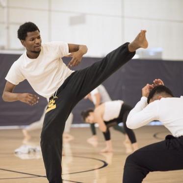 A capoeira student kicks his outstretched leg over another crouching student.