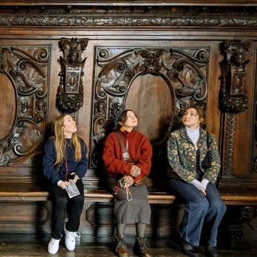 3 students sit on a bench looking up against an intricately carved wood wall.