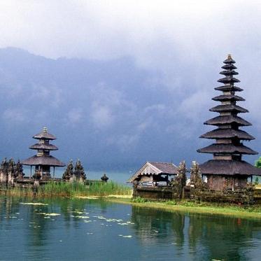 Photo of a small outcropping in water with a large and a small temple in Bali with mist and mountains in the backdrop.